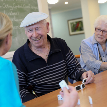Elderly man smiling while playing dominoes with two women at a table in a communal setting.